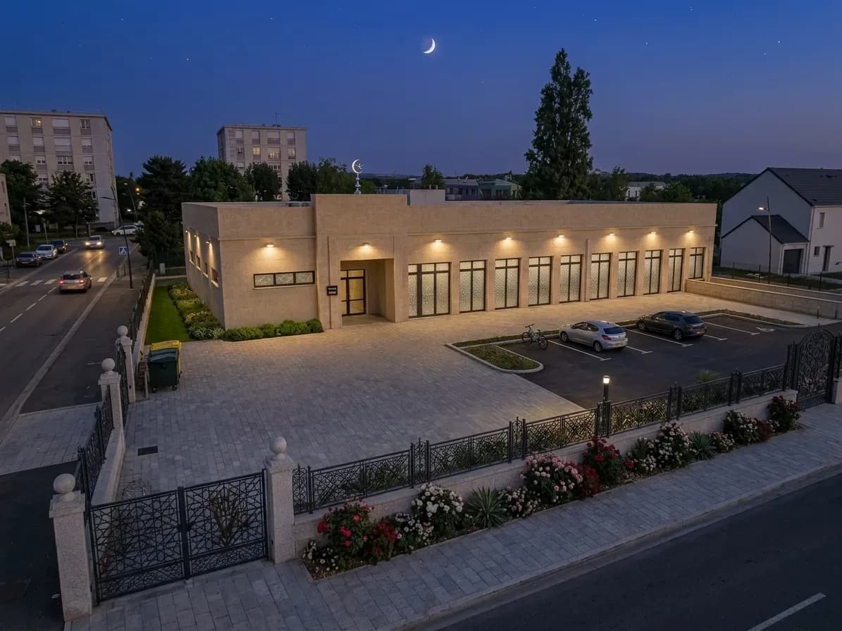Vue de projet aérienne : la mosquée à la tombée du jour, façade en pierre, parvis pavé, jardin fleuri et parking, tels que prévus en phase 2